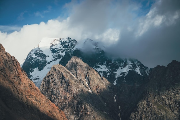 Malerische Landschaft mit großen Felsen und schneebedeckten Bergen im Sonnenlicht in niedrigen Wolken. Wunderbare Aussicht auf den Berggipfel mit Schnee bei Sonnenschein bei bewölktem Himmel. Fantastische Landschaft mit sonnenbeschienenem schneeweißen Gipfel.