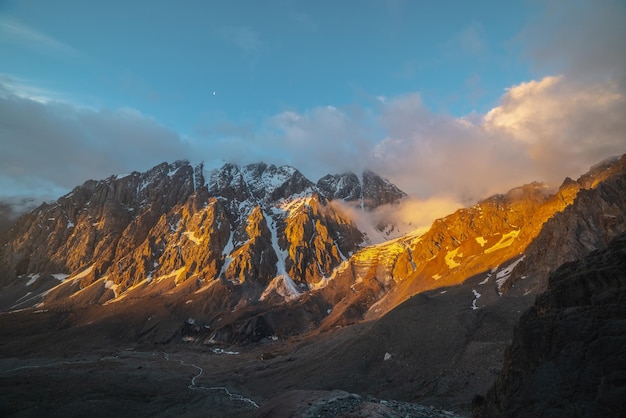 Malerische Landschaft mit schneebedeckter Bergwand und vertikalem Gletscher mit Eisfall in niedrigen Wolken in goldenen Sonnenaufgangsfarben Bunte Aussicht auf schneebedeckte Berge und Felsen in goldenem Morgensonnenlicht in niedrigen Wolken