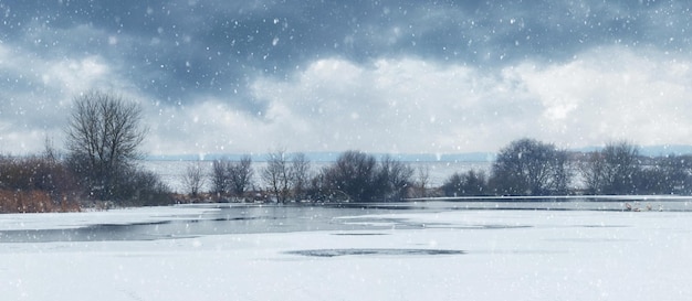 Malerische Winterlandschaft mit Schneefall am Fluss und bewölktem Himmel