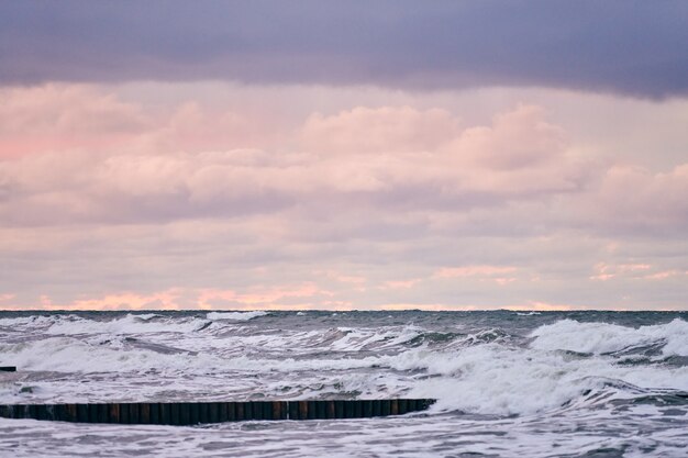 Malerischer Blick auf lila bewölkten Himmel und Meer mit schäumenden Wellen. Vintage lange hölzerne Wellenbrecher, die sich weit ins Meer erstrecken. Windiges Wetter, schöne abendliche Ostseelandschaft.