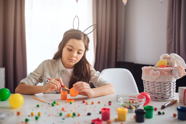 Malestar adolescente se prepara para la Pascua solo. Se sienta a la mesa y pinta huevos. Chica usar orejas de conejo. Dulces coloridos y decoración en la mesa.