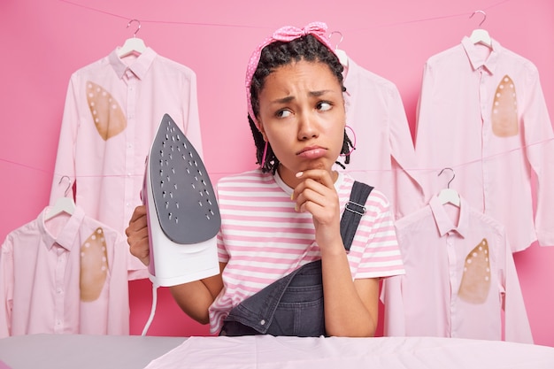 Malestar joven ama de llaves afroamericana con rastas mira tristemente lejos no quiere planchar ropa sostiene plancha eléctrica usa diadema camiseta a rayas y overoles está cansada de las tareas domésticas.