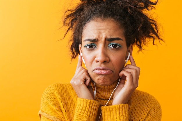 Malestar mujer africana vistiendo suéter que se encuentran aisladas, escuchando música con auriculares