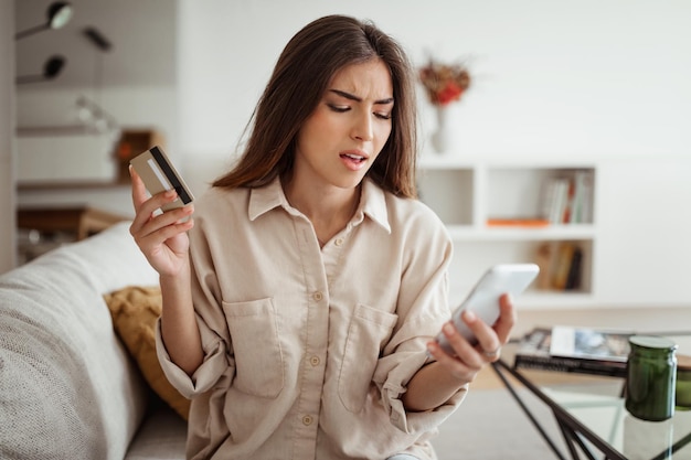 Malestar triste mujer europea milenaria con tarjeta de crédito mirando el teléfono en una sala de estar minimalista