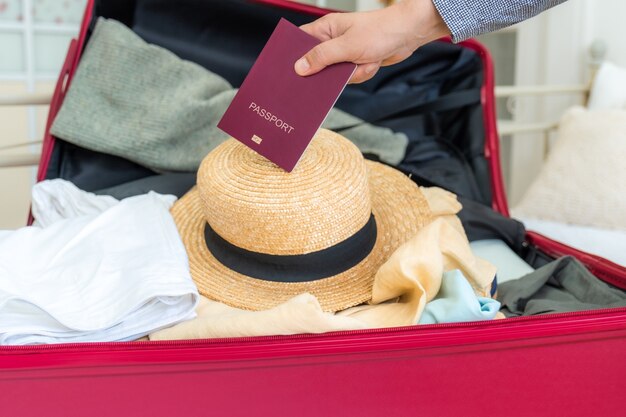 Maleta rosa sobre la cama con ropa, gorro de verano y pasaporte en la mano, lista para viajar.