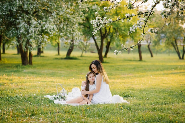 Mamá abraza a su hija sentada en una manta en un jardín florido de primavera Amor materno por un niño