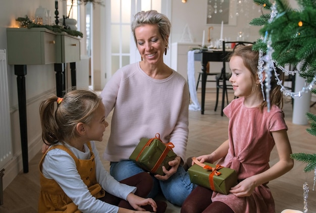 Foto mamá alegre y sus lindas hijas intercambian regalos de navidad sentadas en el suelo cerca del árbol de navidad