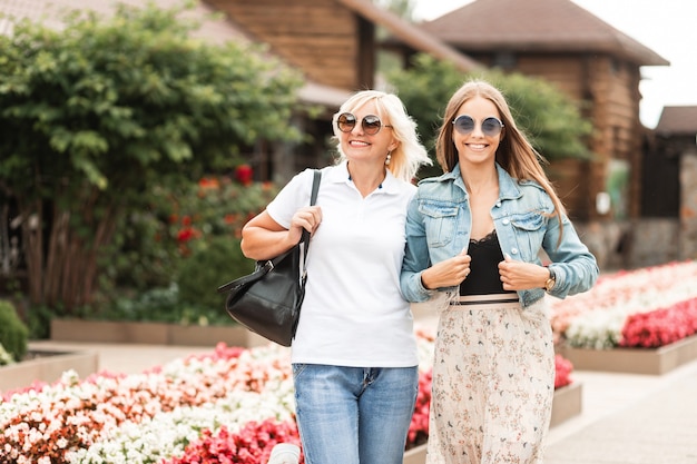 Mamá anciana con estilo en jeans y un polo blanco con una joven hermosa hija sonriente con gafas de sol y una chaqueta de mezclilla caminan juntos y viajan el fin de semana