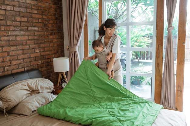 Mamá asiática y su bebé limpiando y arreglando una manta en la cama