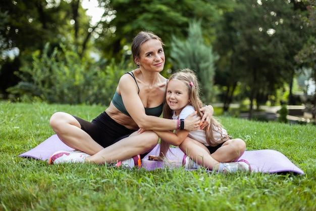Mamá atlética abraza a su hija pequeña mientras hace ejercicio en la alfombra en el parque Familia saludable Cuidado concepto de amor maternidad