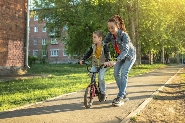 Mamá ayuda a un niño a aprender a andar en bicicleta de dos ruedas en el parque Unas agradables vacaciones deportivas de verano para niños