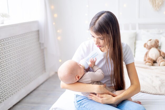 Mamá con un bebé en los brazos alimenta leche de un biberón en la cama en casa en una habitación luminosa el concepto de comida para bebés mamá se preocupa por un niño pequeño