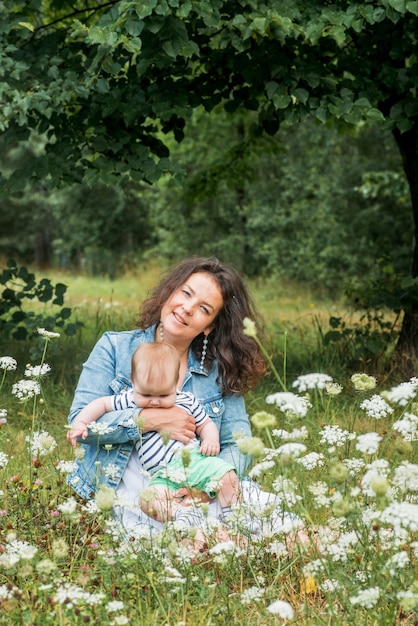 Mamá y bebé están sentados en un parque debajo de un árbol entre las flores y sonriendo. Campo de flores, picnic al aire libre.