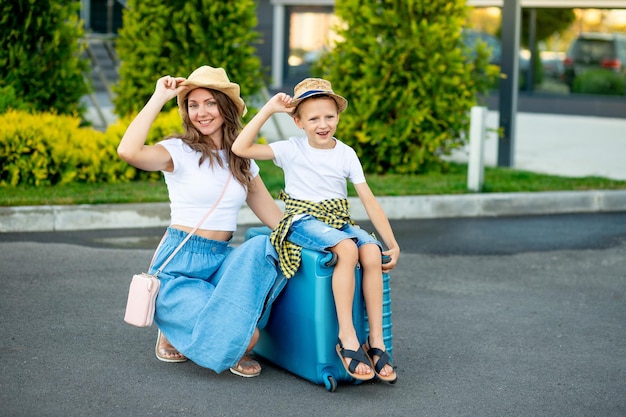 Mamá y bebé felices están sentados en una maleta azul frente al aeropuerto y se van de vacaciones o viajan al aeropuerto