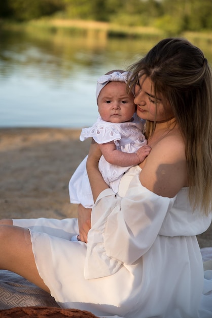 Foto mamá con un bebé recién nacido se sienta en la playa en la arena mamá sostiene a un bebé recién nacido en sus brazos