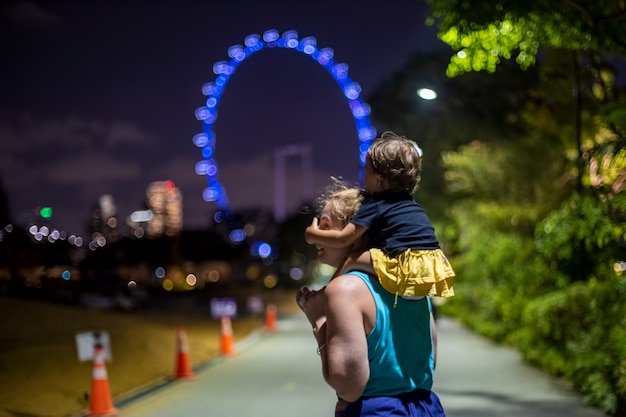 Mamá con un bebé sobre sus hombros en el parque de la ciudad por la noche con el telón de fondo de la noria