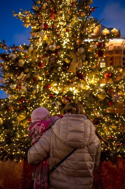 Mamá y bebé en sus brazos miran el árbol de Navidad que brilla intensamente en la vista posterior de la feria de la calle