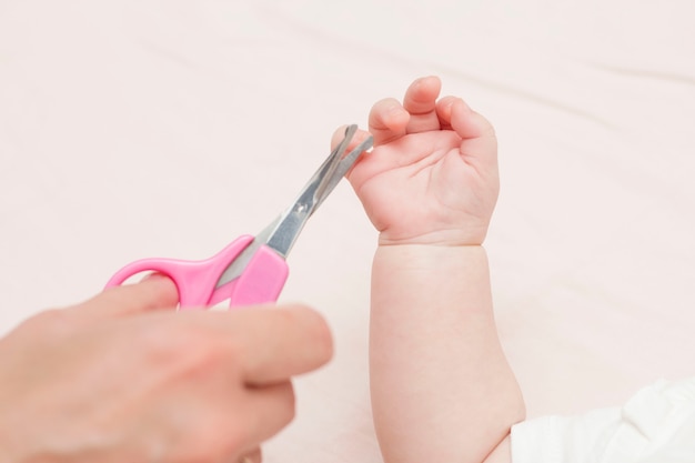Mama bekommt eine Maniküre mit einer Nagelschere und schneidet ihrem süßen Baby die Nägel, während sie auf dem Bett liegt. Das Konzept der Kinderbetreuung.
