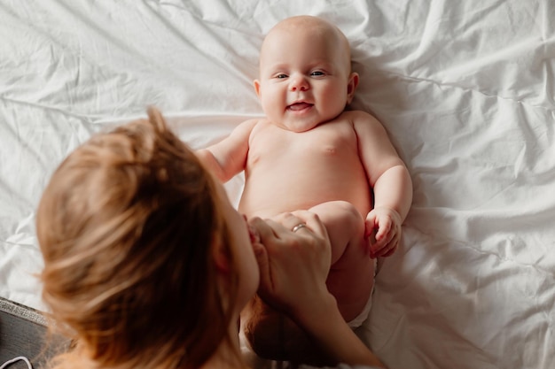 Mamá besa los pies descalzos de un bebé recién nacido sonriente acostado en la cama mujer joven con un bebé en casa