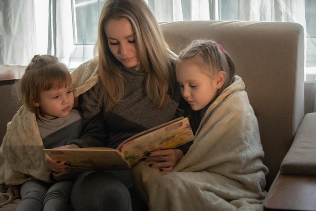 Mamá en casa en el sofá lee un libro a sus hijas pequeñas