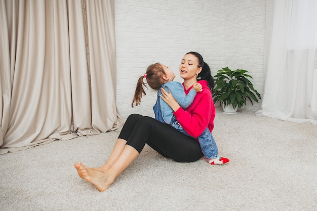 Mamá caucásica sonriente está sentada en el piso de la sala de estar y jugando con su pequeña hija. Feliz madre amorosa diviértete con la niña el fin de semana en casa familiar.