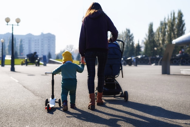 Mamá con cochecito y bebé con scooter caminando juntos en el parque