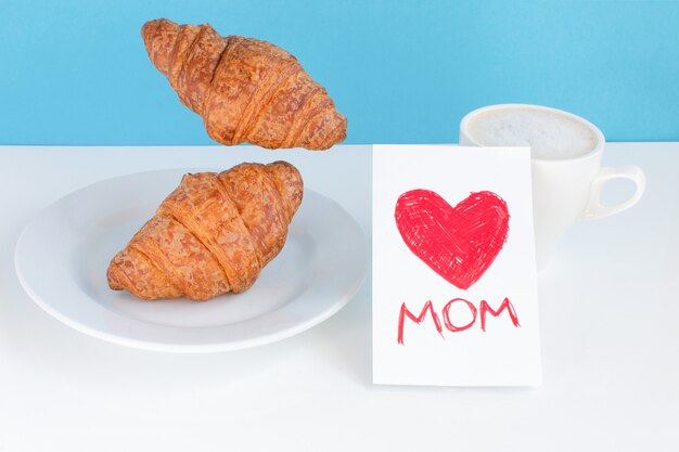 Mamá con un corazón rojo en una tarjeta, una taza blanca y croissants en un plato y volando sobre fondo azul.