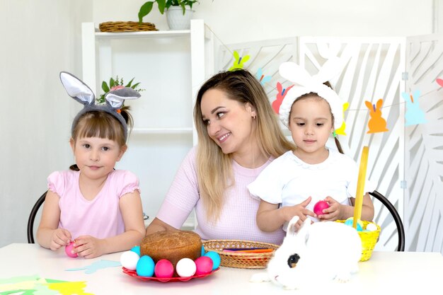 Foto mamá con dos hijas pequeñas se sienta en una mesa blanca con un pastel y una canasta de huevos de pascua