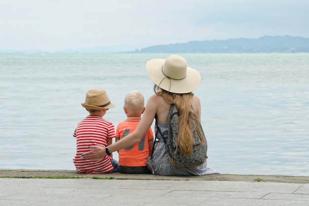 Mamá y dos hijos se sientan en el muelle y admiran el mar y las montañas a lo lejos. Vista trasera