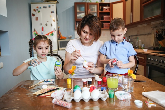 Mamá con dos niños decoran huevos de Pascua sentados a la mesa en casa en la cocina