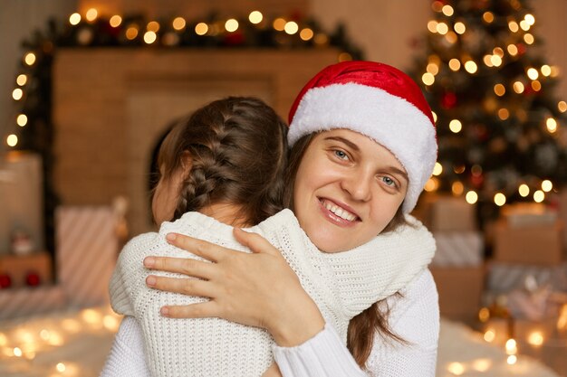 Mamá e hija cerca del árbol de Navidad y la chimenea abrazándose con felicidad