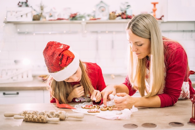 Mamá e hija en la cocina glasean galletas de jengibre en suéteres rojos y gorro de Papá Noel con luces