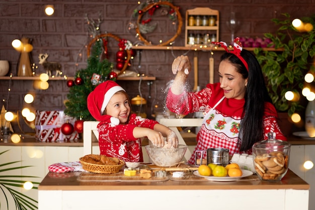 Mamá e hija en una cocina oscura con un árbol de Navidad están preparando galletas de jengibre para Año Nuevo o Navidad, sonriendo y divirtiéndose juntas en anticipación de las vacaciones con un sombrero de Santa Claus