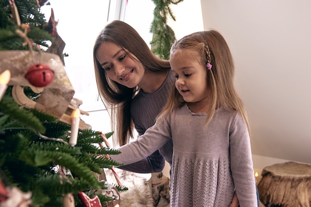 Mamá e hija decoran el árbol de Navidad. Retrato de familia amorosa en Beautiful holdiay DIY decoración del hogar hecha a mano. Acogedora habitación decorada con velas y árbol de Navidad con regalos debajo