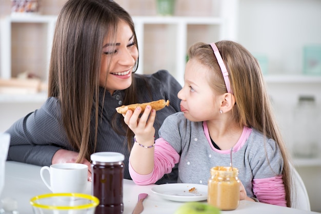 Mamá e hija desayunan en el ambiente de la cocina y hablan. Una niña linda come pan con mantequilla de maní.