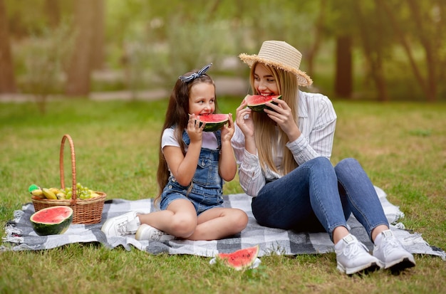 Mamá e hija disfrutando de un picnic familiar comiendo sandía sentada sobre una manta en el hermoso parque verde. Fin de semana con el concepto de madre