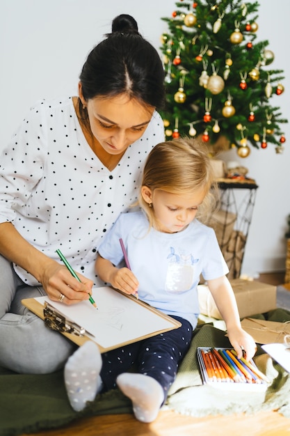 Mamá e hija están escribiendo una carta a Papá Noel en la pared de un árbol de Navidad y regalos Mujer joven enseña a una niña linda a dibujar una tarjeta de Año Nuevo Momentos acogedores familiares