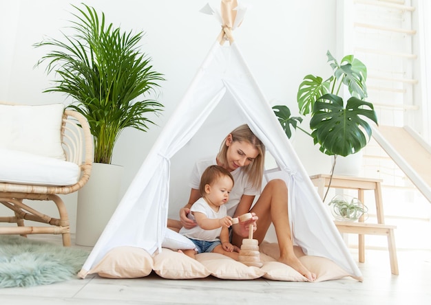 Mamá e hija están jugando con una pirámide en desarrollo mientras se sientan en un wigwam en una habitación de luz minimalista moderna con plantas de la casa