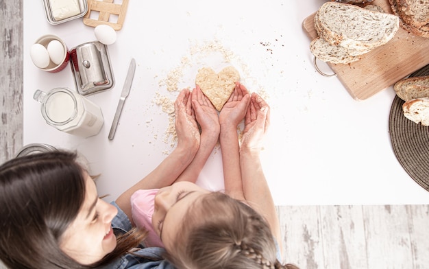 Mamá e hija están preparando galletas en forma de corazón en una gran mesa de cocina blanca.