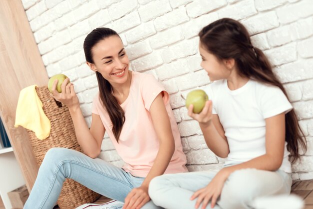 Mamá e hija están sentadas en el piso y comiendo una manzana.