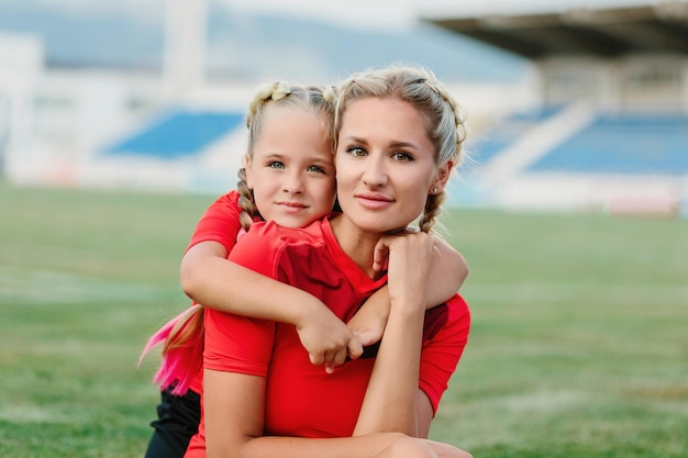 Mamá e hija están sentadas con ropa deportiva en el campo de fútbol y abrazándose