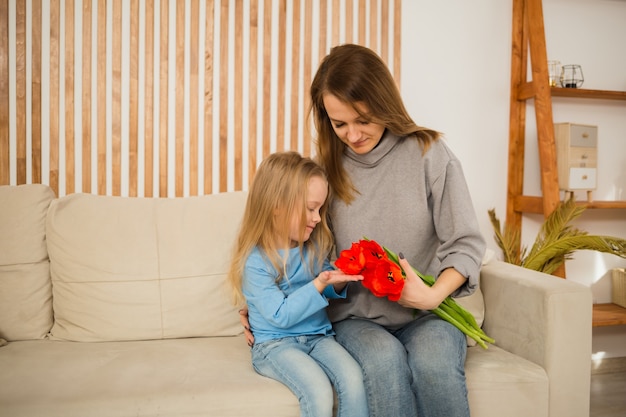 Foto mamá e hija están sentadas en el sofá y mirando un ramo de tulipanes rojos en la habitación