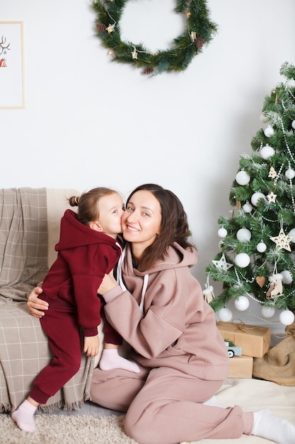 Mamá e hija europea de ojos azules jugando junto a un árbol de Navidad decorado