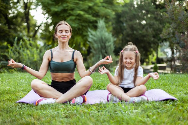 Mamá e hija meditan practicando yoga juntos en la alfombra en el parque Maternidad Pasando tiempo Mamá Entrenador concepto de familia saludable