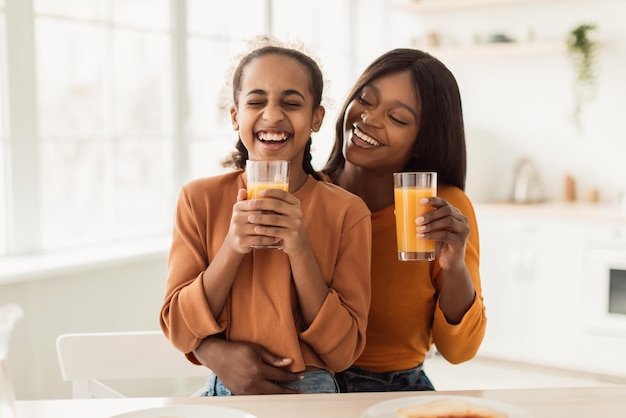 Mamá e hija negras bebiendo jugo de naranja riéndose en la cocina