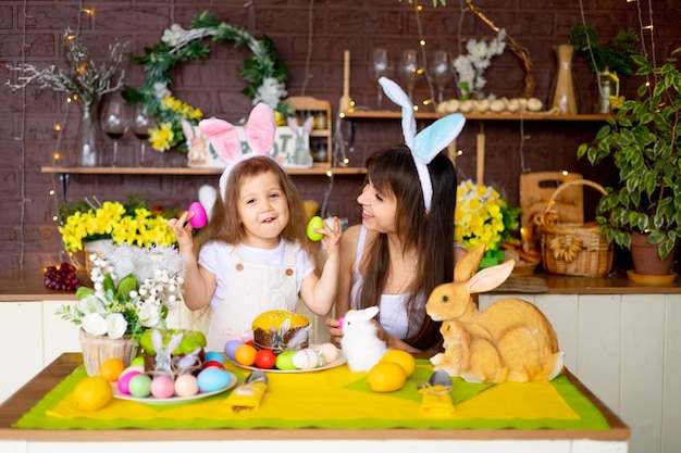 Mamá e hija de Pascua con huevos de Pascua y pastel en la mesa festiva están sonriendo y preparándose para la Pascua en casa en la cocina en las orejas de una liebre