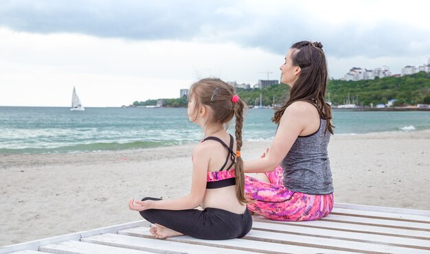 Mamá e hija en ropa deportiva practican yoga en la orilla del mar. Valores familiares y estilo de vida saludable.