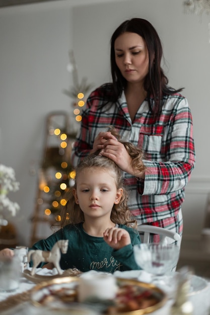 Mamá e hija con ropa navideña de moda se sientan en la mesa festiva y se peinan en Nochebuena