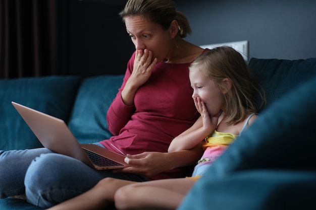 Mamá e hija sorprendidas están sentadas en el sofá y mirando la pantalla del portátil