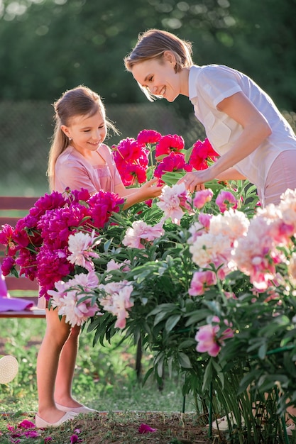 Mamá e hija con un vestido rosa de lino cuidan peonías en el jardín, riegan las flores. Sonríe y ríe perfectamente. Iluminado por los rayos del sol poniente de la tarde.